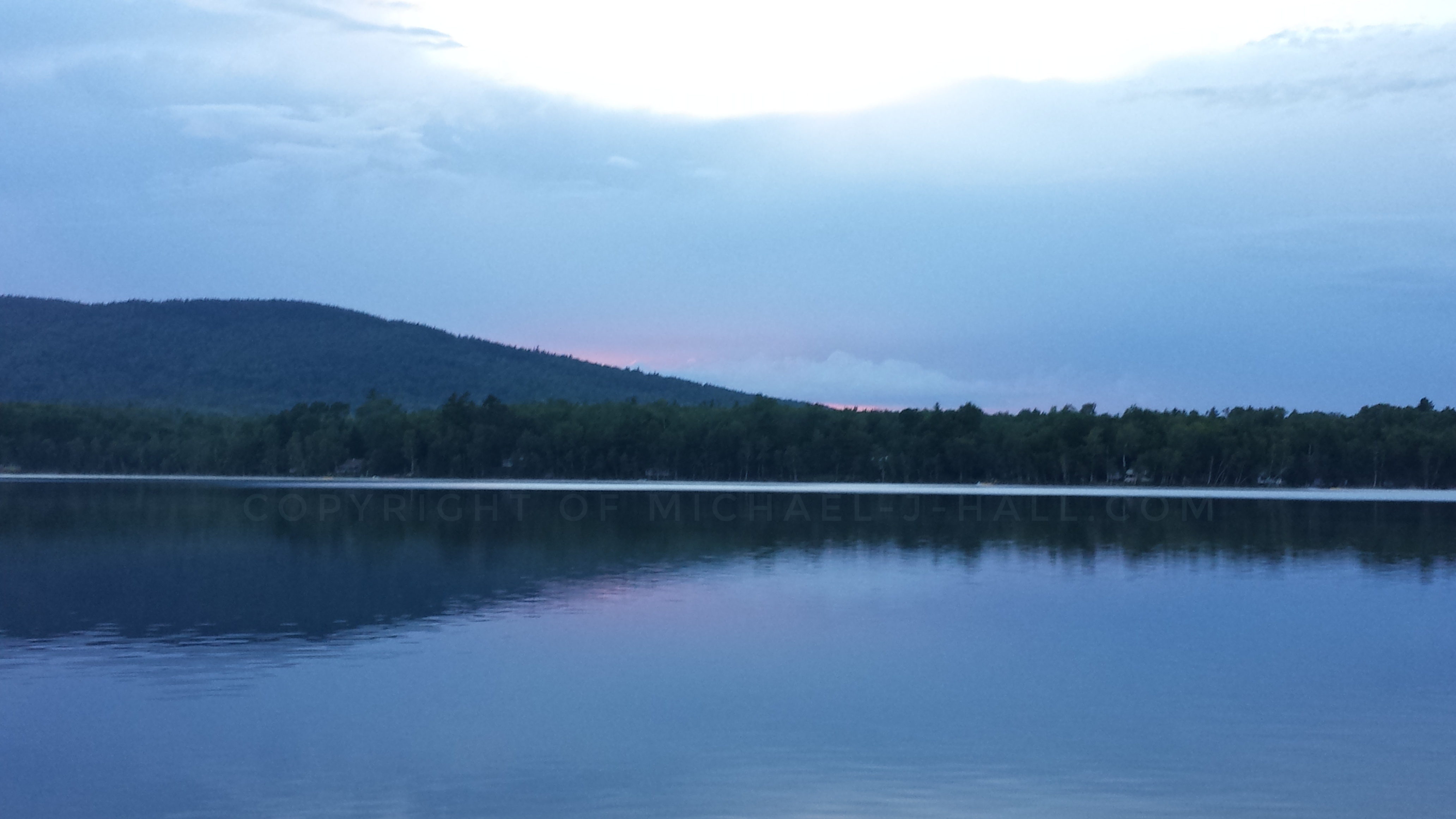 Dusky serenity at the water's edge with sun settling behind distant hills and thin clouds overhead static from the lack of wind making for very still waters...